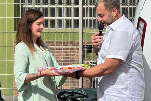 Eine Frau in einem hellen Kleid überreicht einem Mann mit Mikrofon einen Teller mit Essen vor einem weißen Van.