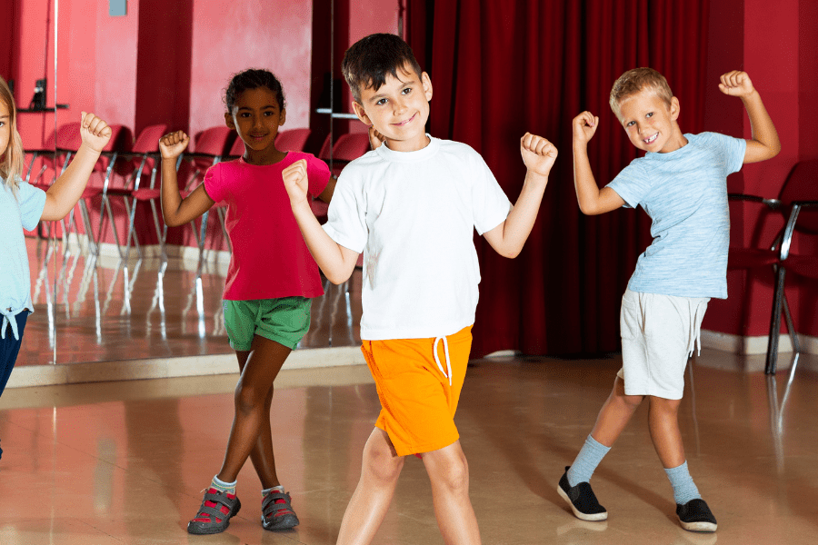 Sechs Kinder in sportlicher Kleidung posieren fröhlich in einem Tanzraum mit Spiegeln und roten Wänden.