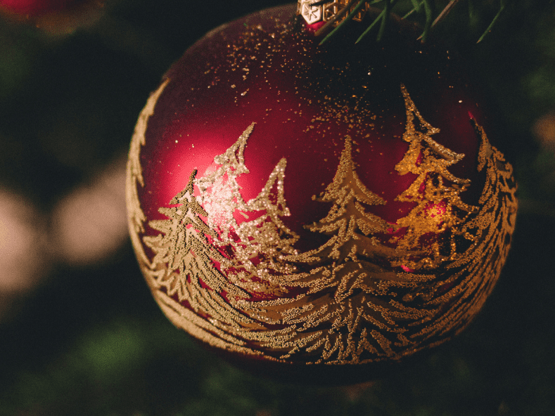Rote Weihnachtskugel mit goldener Baumgrafik h&auml;ngt an einem Tannenbaum, im Hintergrund verschwommenes Licht.