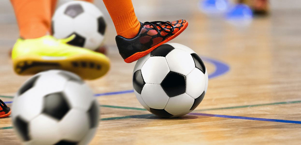 Fußballprofile an einer Sporthalle, zwei Spieler in orangefarbener Socke und verschiedenen Schuhen stehen um einen Ball.