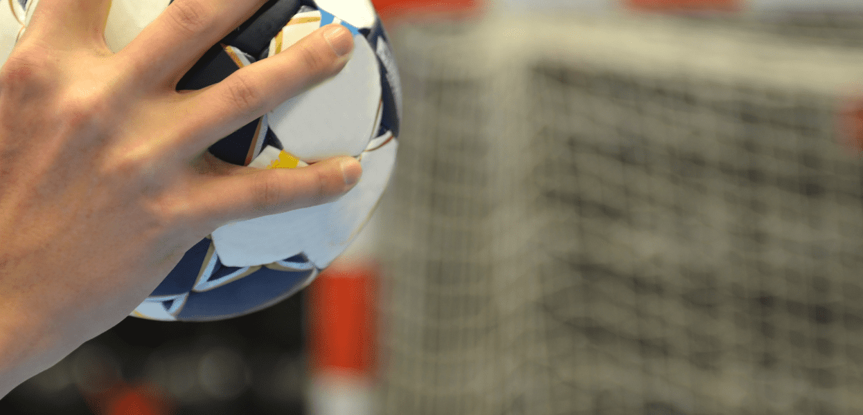 Hand h&auml;lt einen blauen und wei&szlig;en Handball, mit Tor im Hintergrund.