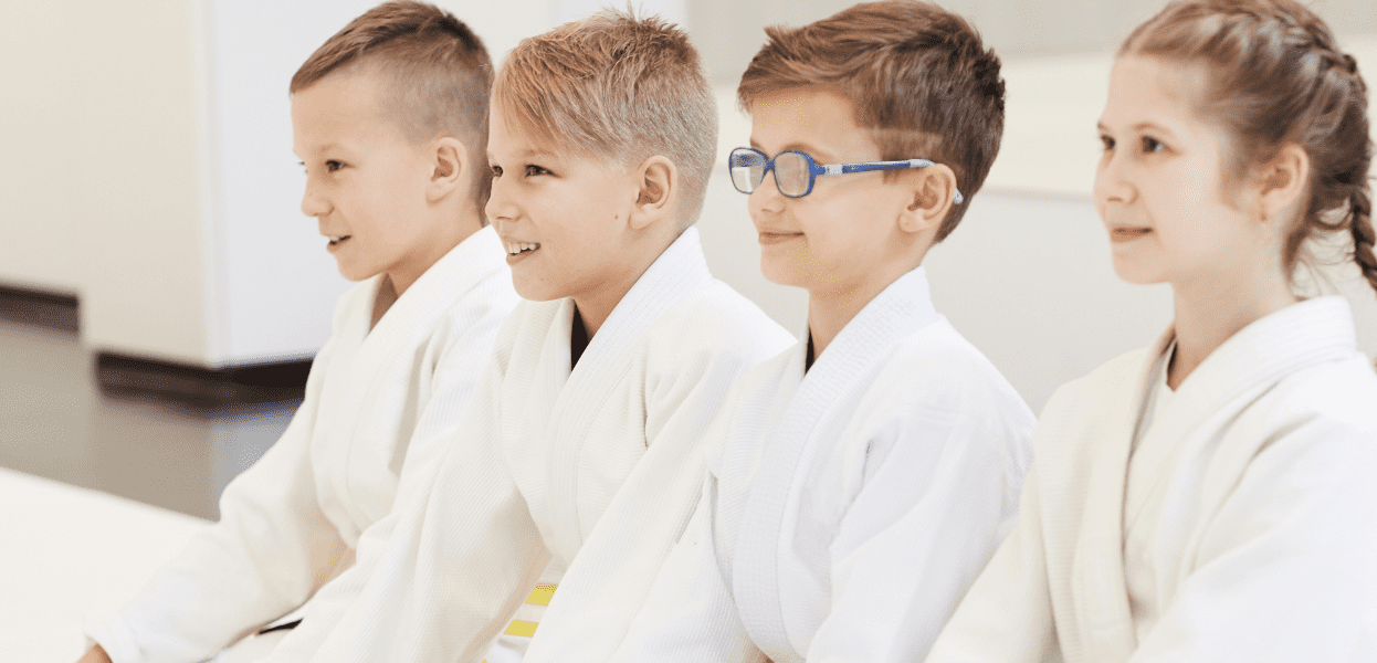Vier Kinder in weißen Judokis warten auf der Matte, Blick nach vorne gerichtet, lächelnd. Helle, moderne Umgebung.