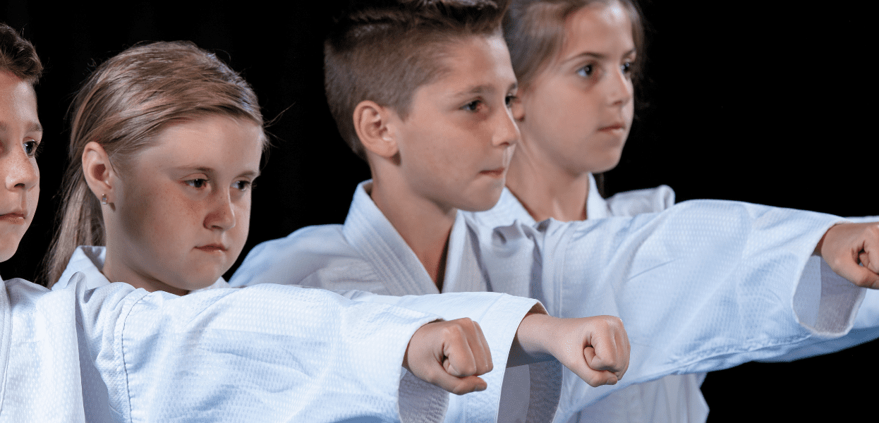 Vier Kinder in weißen Karateanzügen zeigen Kampftechniken mit erhobenen Fäusten vor schwarzem Hintergrund.