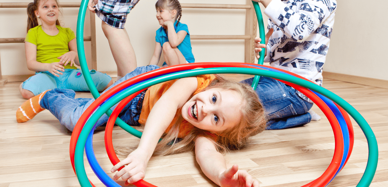 Vier Kinder spielen in einem Raum mit Hula-Hoop-Reifen, zwei Kinder liegen auf dem Boden und lächeln.