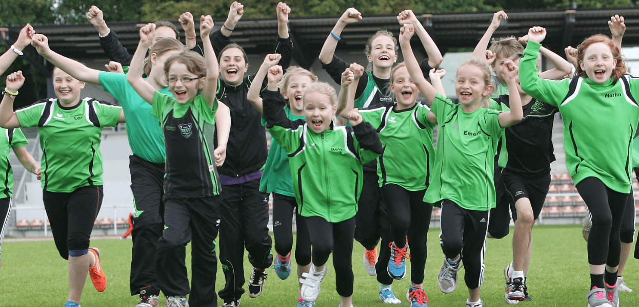 Gruppe von Kindern in gr&uuml;nen Trikots jubelt und l&auml;uft fr&ouml;hlich &uuml;ber einen Sportplatz.