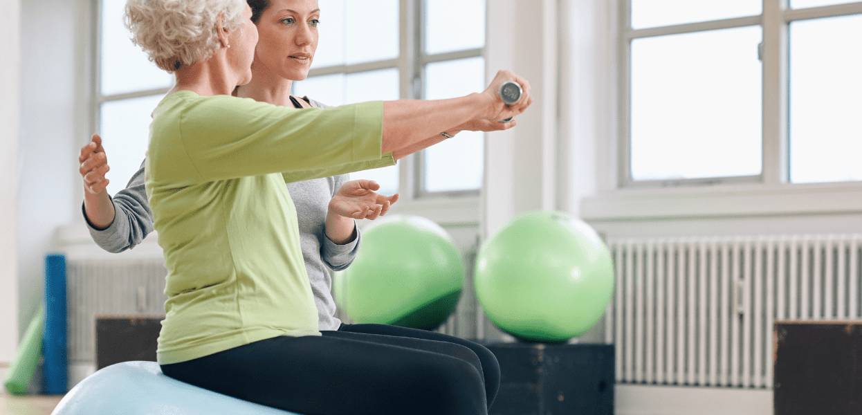 Zwei Frauen, eine &auml;ltere und eine j&uuml;ngere, &uuml;ben gemeinsam mit einem Gymnastikball in einem hellen Trainingsraum.