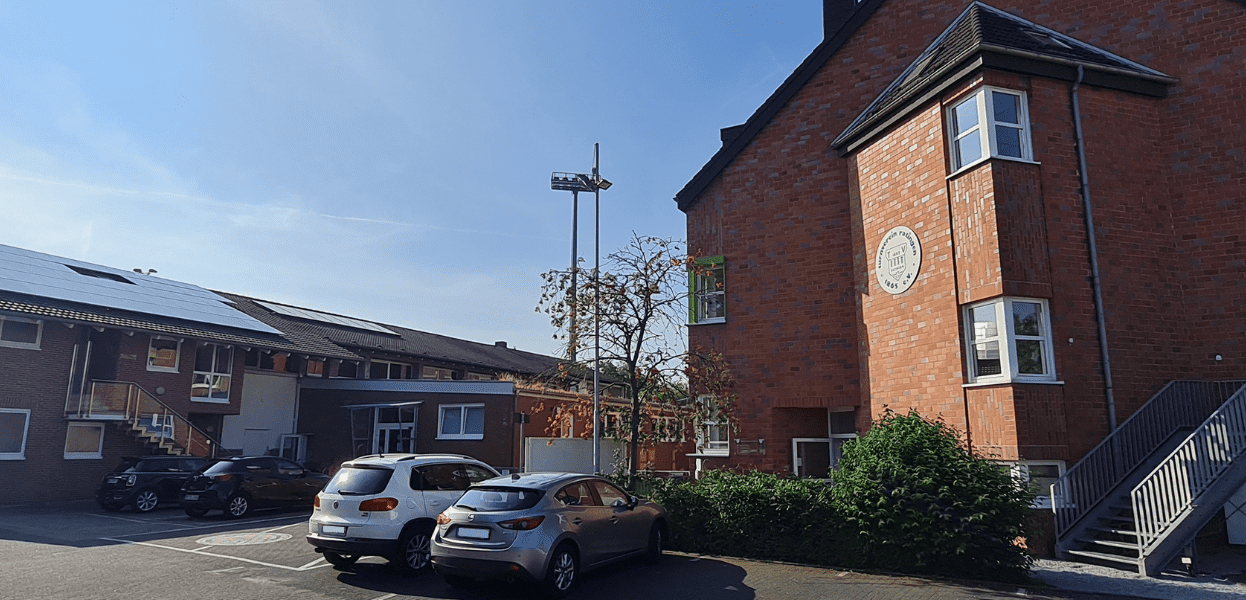 Zwei Geb&auml;ude in einer ruhigen Umgebung, einer mit Solarpanelen, mehrere Autos auf einem Parkplatz.