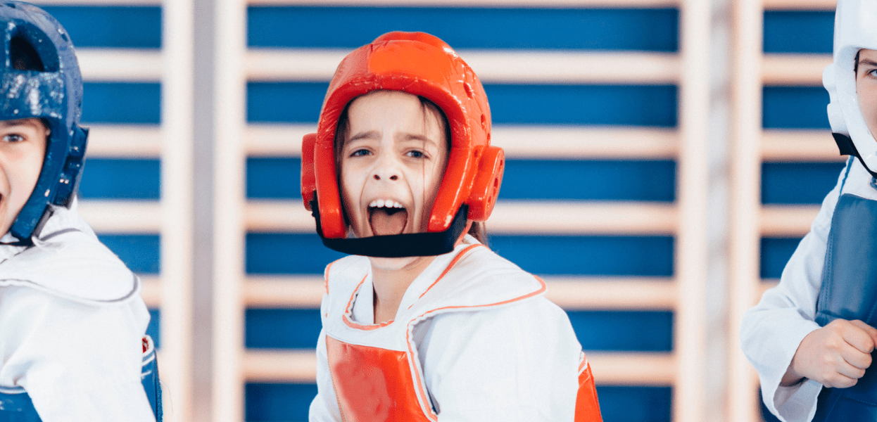 Drei Kinder in Kampfsportausrüstung mit Helmen in verschiedenen Farben posieren energisch vor einem blauen Hintergrund.