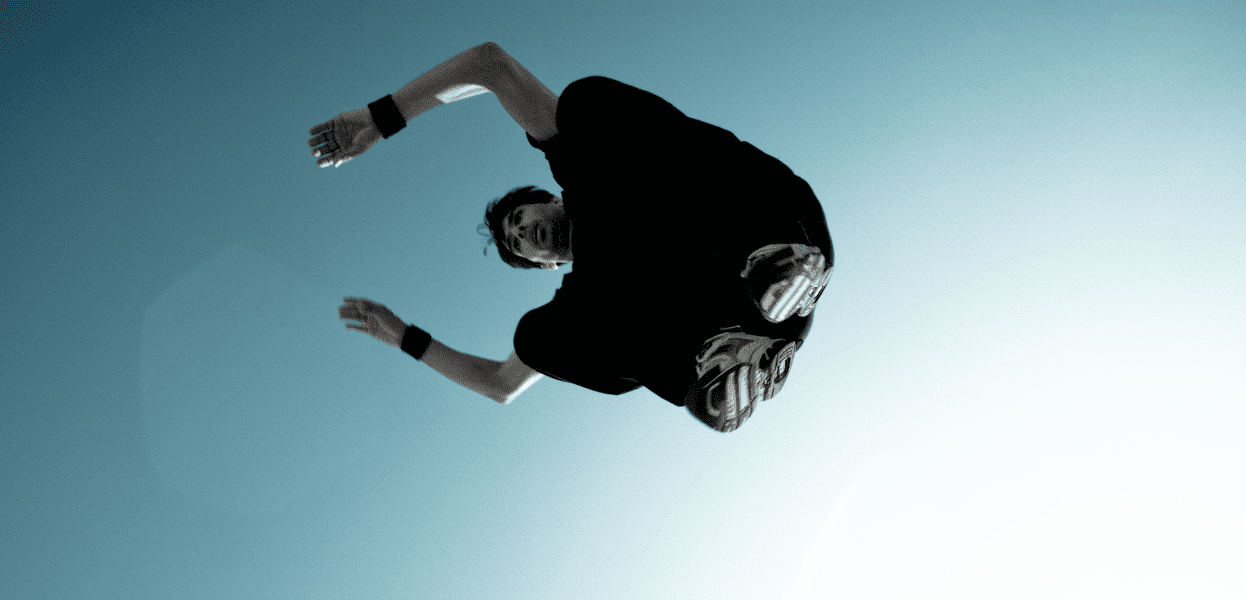 Sprung eines Sportlers mit ausgestreckten Armen vor hellem Himmel, w&auml;hrend er sich in der Luft dreht.