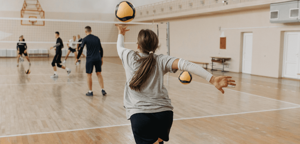 Eine Spielerin wirft einen Volleyball in einer Sporthalle, während andere Spieler im Hintergrund aktiv sind.