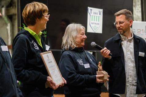 Vier Personen stehen auf einer Veranstaltung, eine h&auml;lt eine Auszeichnung. Im Hintergrund ein Vereinslogo.