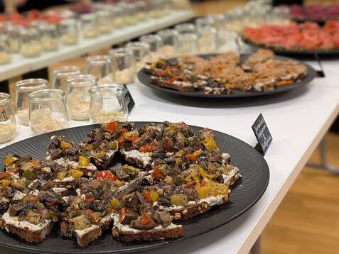 Auf einem Tisch stehen mehrere Platten mit belegten Broten, umgeben von Glasbeh&auml;ltern mit Zutaten.