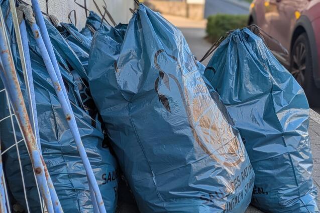 Blaue M&uuml;lls&auml;cke stehen neben einem Gehweg, w&auml;hrend ein Auto und ein Geb&auml;ude im Hintergrund sichtbar sind.