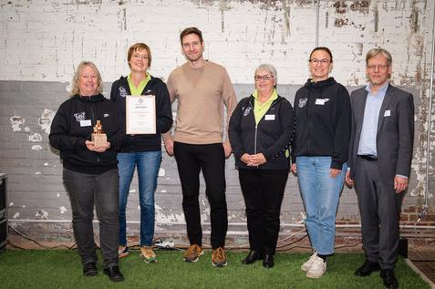 Gruppe von f&uuml;nf Personen steht vor einer Wand, eine h&auml;lt eine Urkunde f&uuml;r den ersten Platz und eine Troph&auml;e in der Hand.