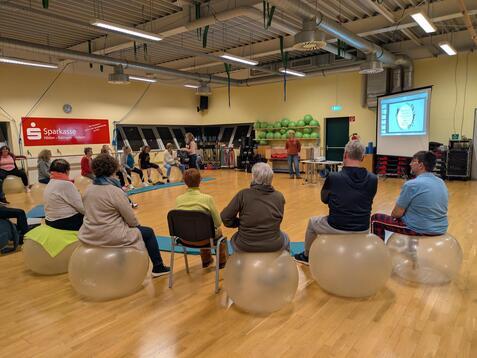 Teilnehmer sitzen auf Gymnastikb&auml;llen in einem Kursraum, w&auml;hrend eine Dozentin eine Pr&auml;sentation leitet.