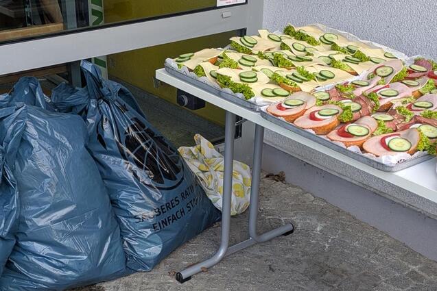 Tisch mit belegten Br&ouml;tchen und gr&uuml;nen Gem&uuml;sebel&auml;gen neben gro&szlig;en blauen S&auml;cken auf einem Pflasterboden.