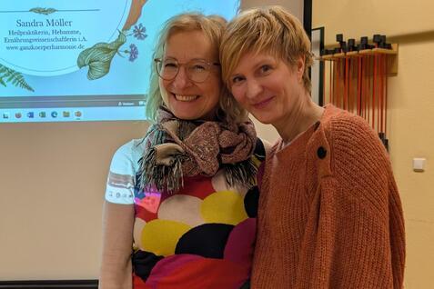 Zwei Frauen stehen l&auml;chelnd nebeneinander vor einem Pr&auml;sentationsbildschirm mit dem Titel &bdquo;N&auml;hrstoffe mit Achtsamkeit&ldquo;.