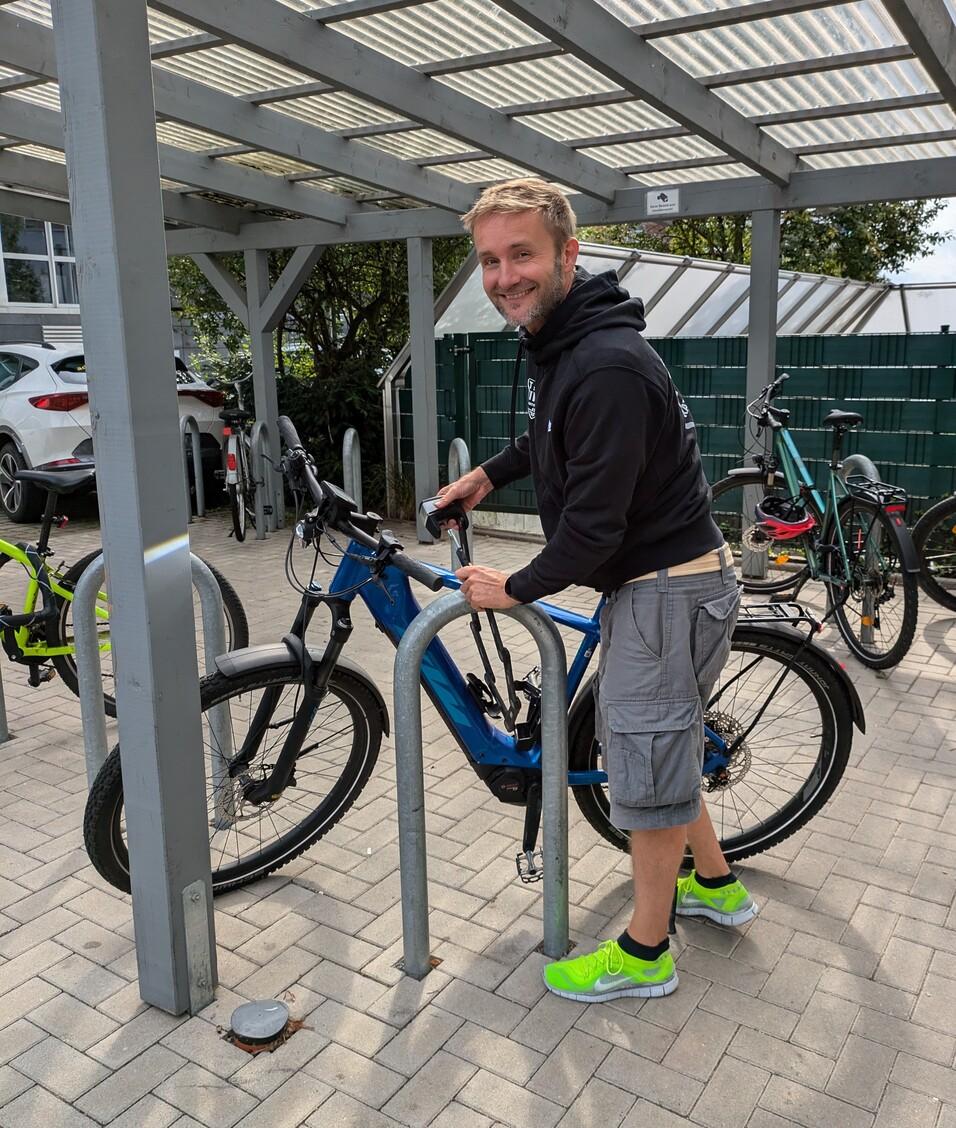 Mann mit kurzem Haar und grünen Schuhen steht neben einem blauen E-Bike in einem Fahrradparkbereich.