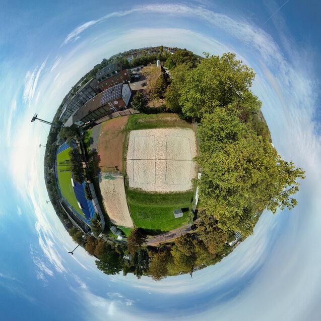 Luftaufnahme eines Sportgeländes mit Sandplätzen, Rasenflächen und angrenzenden Gebäuden unter blauem Himmel.