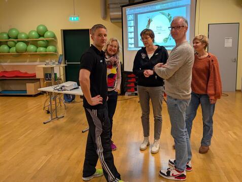 Sechs Personen stehen in einem Raum, diskutieren, w&auml;hrend eine Pr&auml;sentation im Hintergrund l&auml;uft.