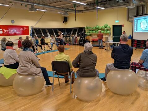 Teilnehmer sitzen auf Gymnastikb&auml;llen in einem Kursraum, w&auml;hrend eine Dozentin eine Pr&auml;sentation leitet.