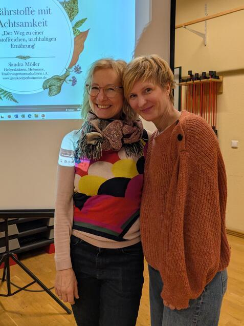 Zwei Frauen stehen l&auml;chelnd nebeneinander vor einem Pr&auml;sentationsbildschirm mit dem Titel &bdquo;N&auml;hrstoffe mit Achtsamkeit&ldquo;.