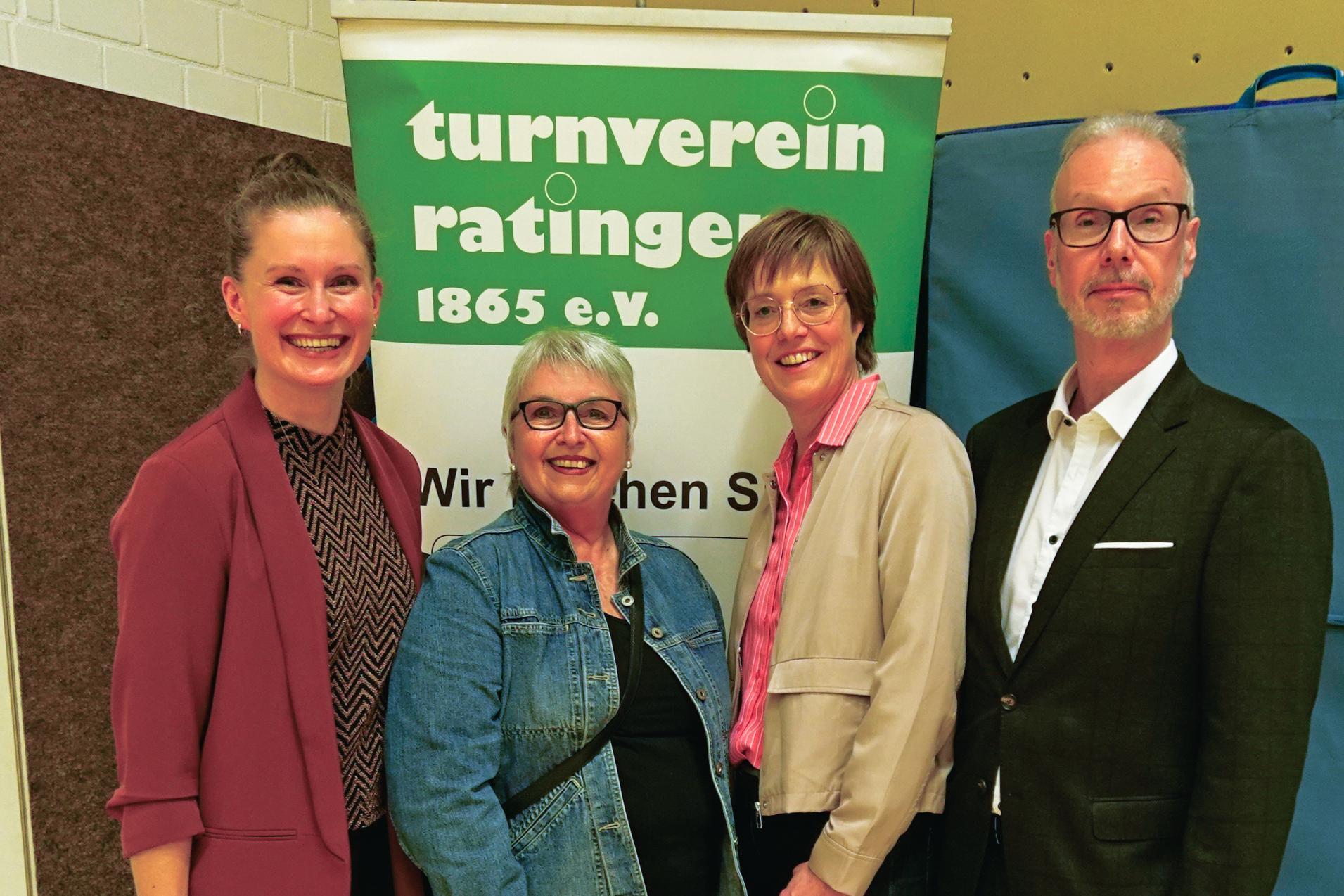 Vier Personen posieren vor einem Banner des Turnvereins Ratingen in einer Sporthalle. Holzfu&szlig;boden im Hintergrund.