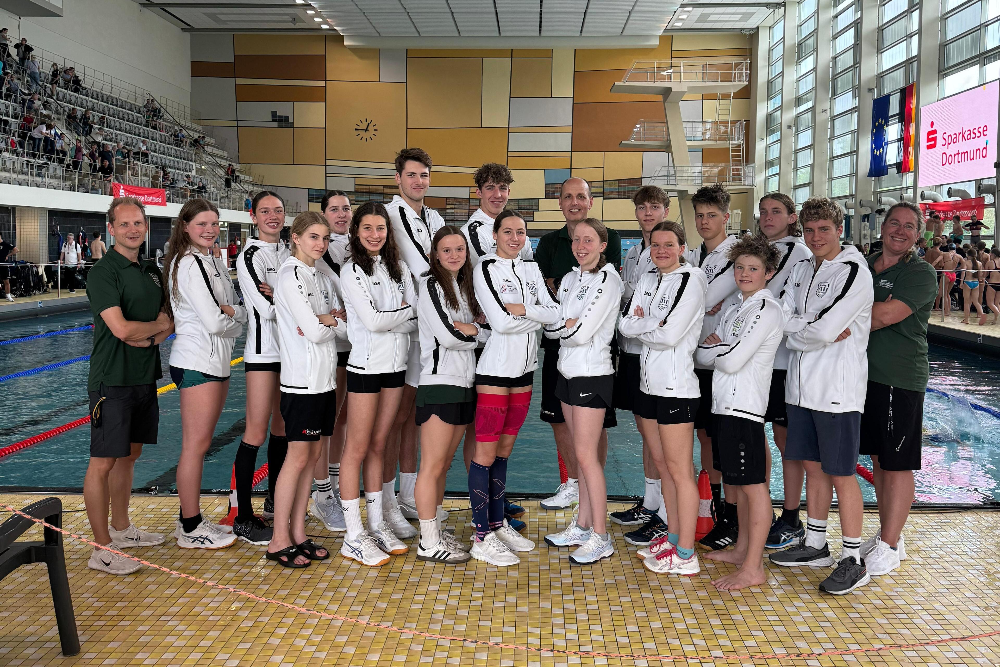 Gruppe von Schwimmern in wei&szlig;en Trainingsjacken posiert am Rand eines Schwimmbeckens in einer Sporthalle.