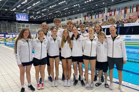 Gruppe junger Schwimmer in wei&szlig;en Trainingsjacken vor einer Schwimmbeckenauflage bei einem Wettkampf.