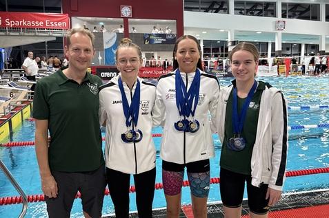 Vier Schwimmerinnen stehen zusammen am Beckenrand, tragen Medaillen und Teamjacken, l&auml;cheln stolz in die Kamera.