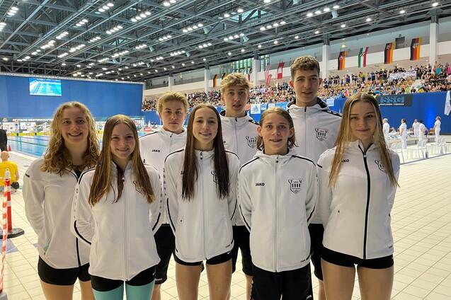 Gruppe junger Schwimmer in wei&szlig;en Trainingsjacken, l&auml;chelnd vor einer Schwimmhalle w&auml;hrend einer Meisterschaft.
