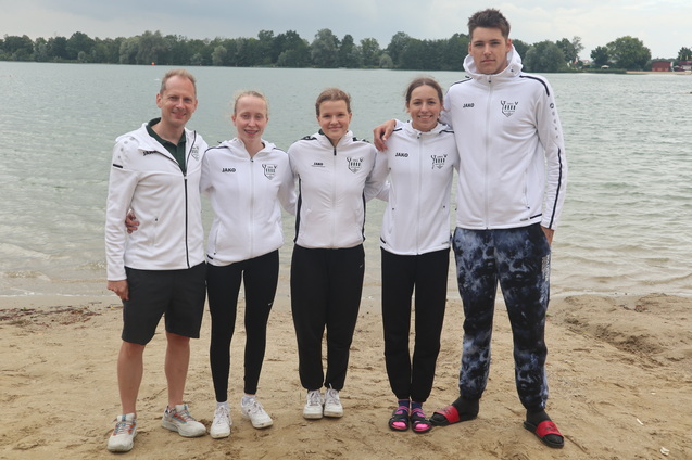 F&uuml;nf Sportler in Trainingsjacken stehen am Sandstrand eines Sees und posieren f&uuml;r ein Gruppenfoto.