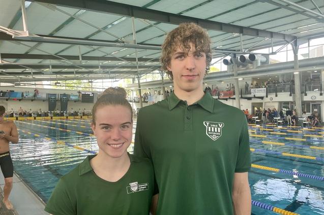 Zwei junge Schwimmer in gr&uuml;nen Vereins-T-Shirts stehen am Beckenrand eines Schwimmbads mit Wasser und Zuschauertrib&uuml;ne.