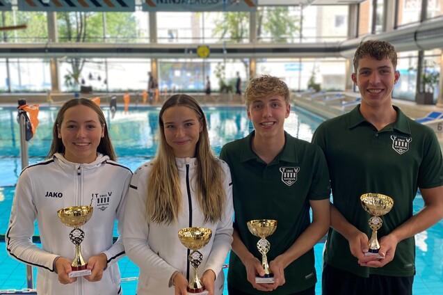 Vier junge Schwimmer halten Pokale in einem Hallenbad mit einer blauen Wasseroberfl&auml;che im Hintergrund.