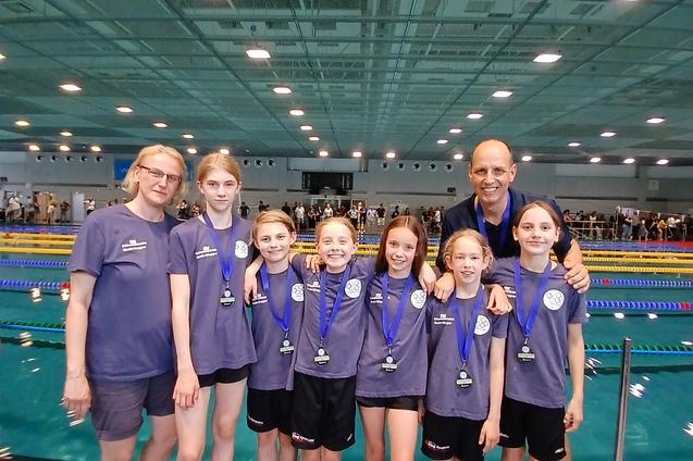 Gruppe von acht Schwimmerinnen und Trainern mit Medaillen in einer Schwimmhalle, im Hintergrund ein Schwimmbecken.