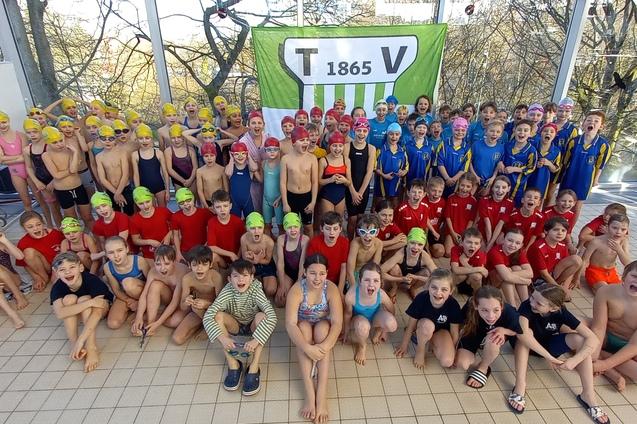 Gruppenfoto von Schwimmern in Badesachen mit unterschiedlichen Badekappen, vor einem Banner des Vereins T 1865.