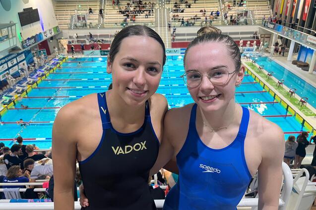 Zwei Schwimmerinnen in Wettkampfanz&uuml;gen stehen neben einem Schwimmbecken, Zuschauer im Hintergrund.