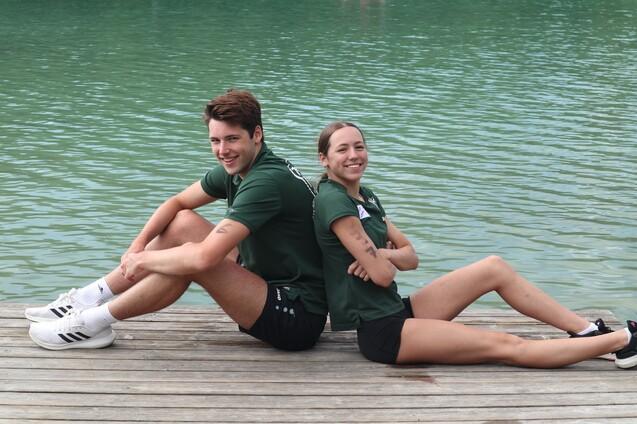 Zwei Sportler sitzen r&uuml;ckw&auml;rts an einem Holzsteg mit Wasser im Hintergrund, beide l&auml;cheln in die Kamera.