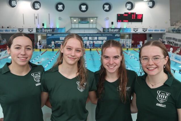 Vier Schwimmerinnen in gr&uuml;nen T-Shirts l&auml;cheln vor einer Schwimmhalle mit Wasserbecken und Zeitnahme-Anzeige.