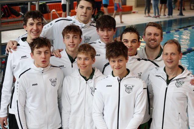 Gruppe von Schwimmern in wei&szlig;en Trainingsjacken, l&auml;chelnd vor einem Schwimmbecken, im Hintergrund weitere Schwimmer.