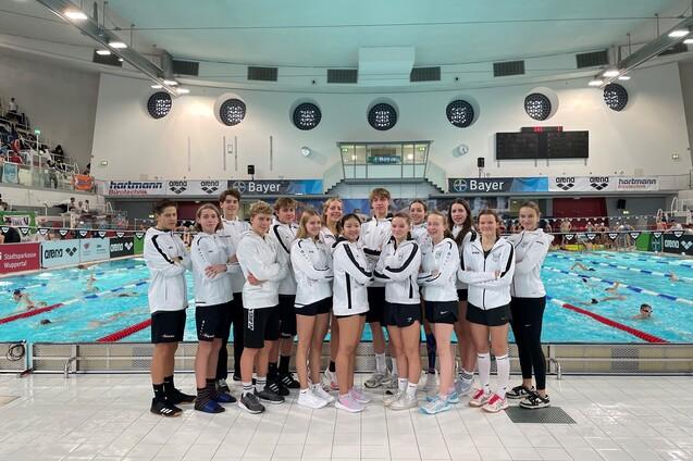 Gruppe von 14 Schwimmern in wei&szlig;en Trainingsjacken vor einem Schwimmbecken mit anderen Schwimmern im Hintergrund.