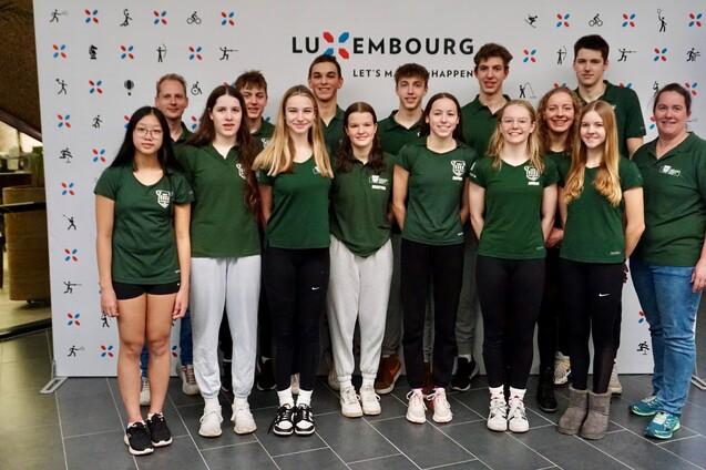 Gruppe von Sportlern in gr&uuml;nen T-Shirts vor einem Banner mit dem Schriftzug &bdquo;LUXEMBOURG &ndash; LET'S MAKE IT HAPPEN&ldquo;.