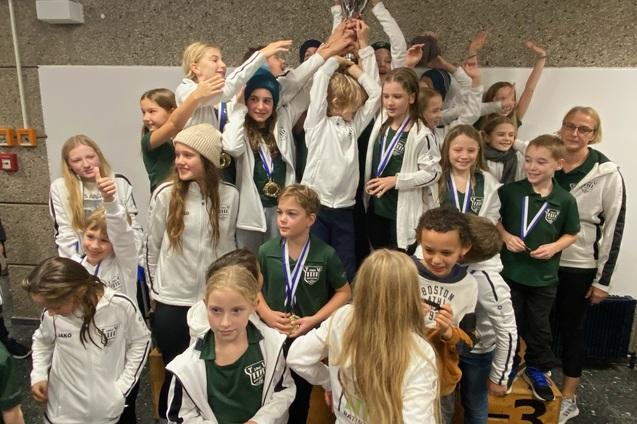 Gruppe von Kindern in Sportjacken feiert mit einem Pokal und Medaillen auf einem Siegertreppchen.