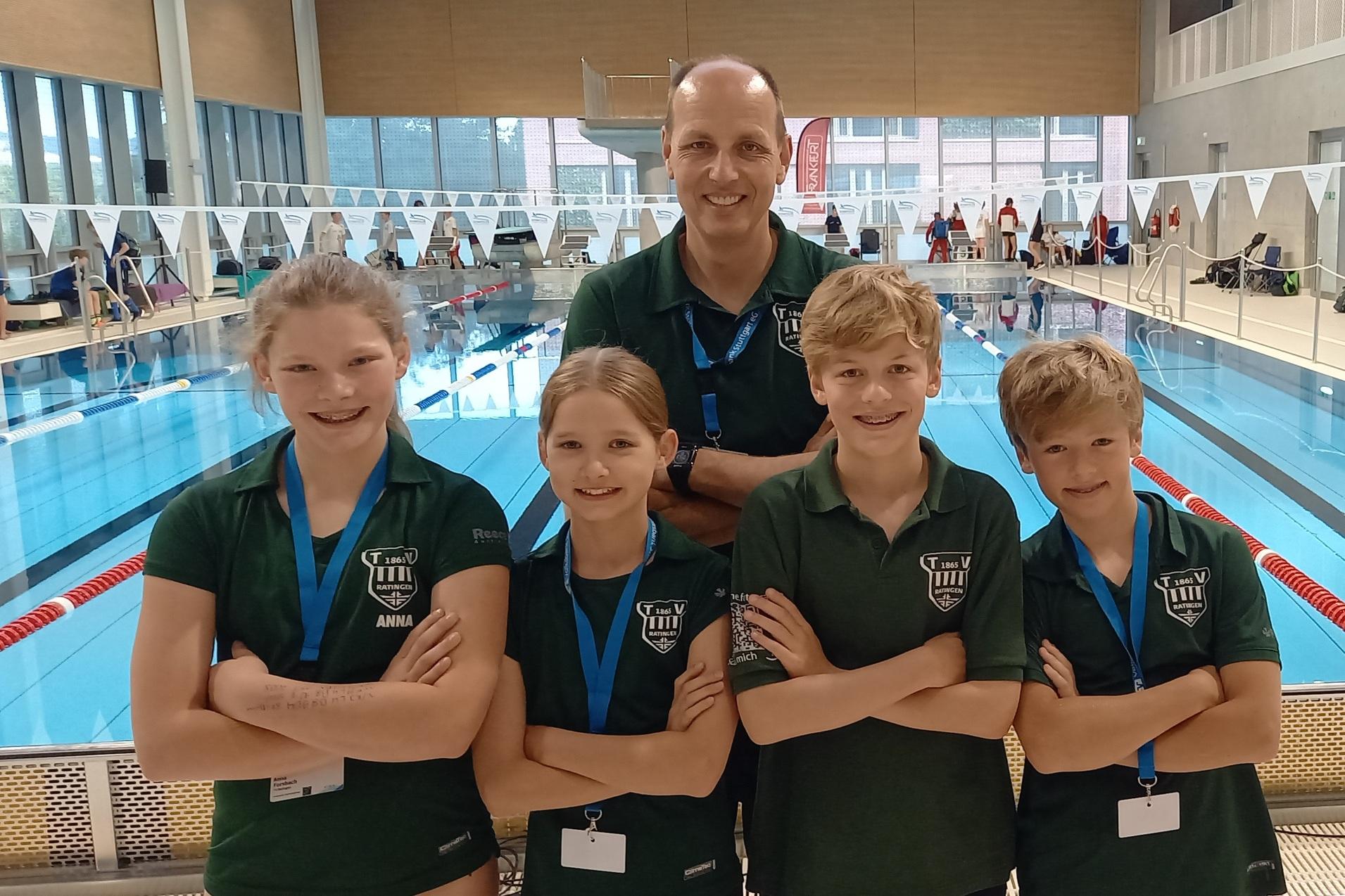 Gruppenfoto mit einem Trainer und drei Schwimmern in grünen T-Shirts am Beckenrand eines Schwimmbads.