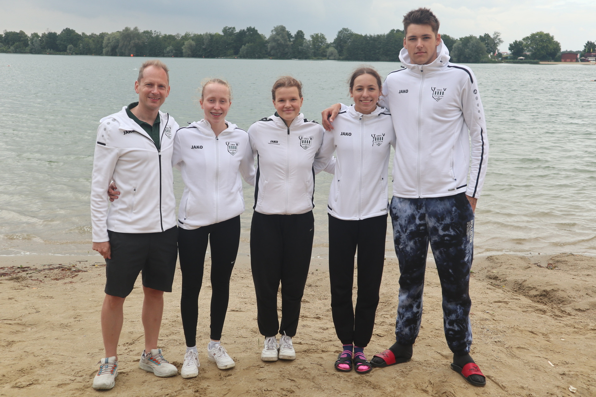 F&uuml;nf Sportler in Trainingsjacken stehen am Sandstrand eines Sees und posieren f&uuml;r ein Gruppenfoto.