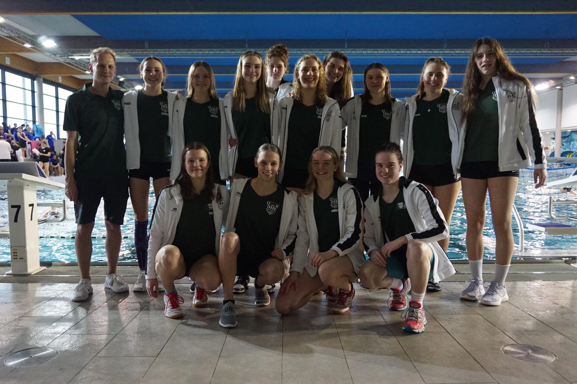 Gruppe von Schwimmerinnen und Trainer am Beckenrand in einer Schwimmhalle, alle tragen Teamjacken und Sportkleidung.