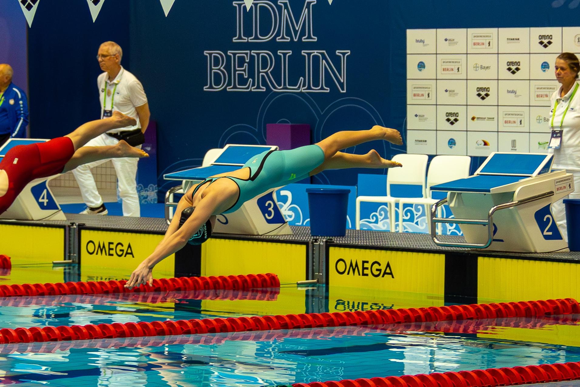 Schwimmerin in elastischem Anzug springt ins Wasser von einem Startblock, w&auml;hrend ein Wettkampf im Hintergrund l&auml;uft.
