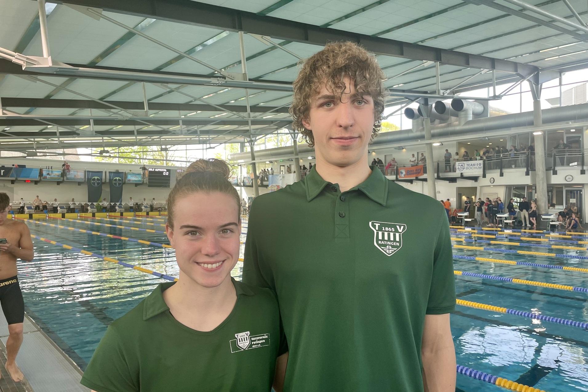 Zwei junge Schwimmer in gr&uuml;nen Vereins-T-Shirts stehen am Beckenrand eines Schwimmbads mit Wasser und Zuschauertrib&uuml;ne.