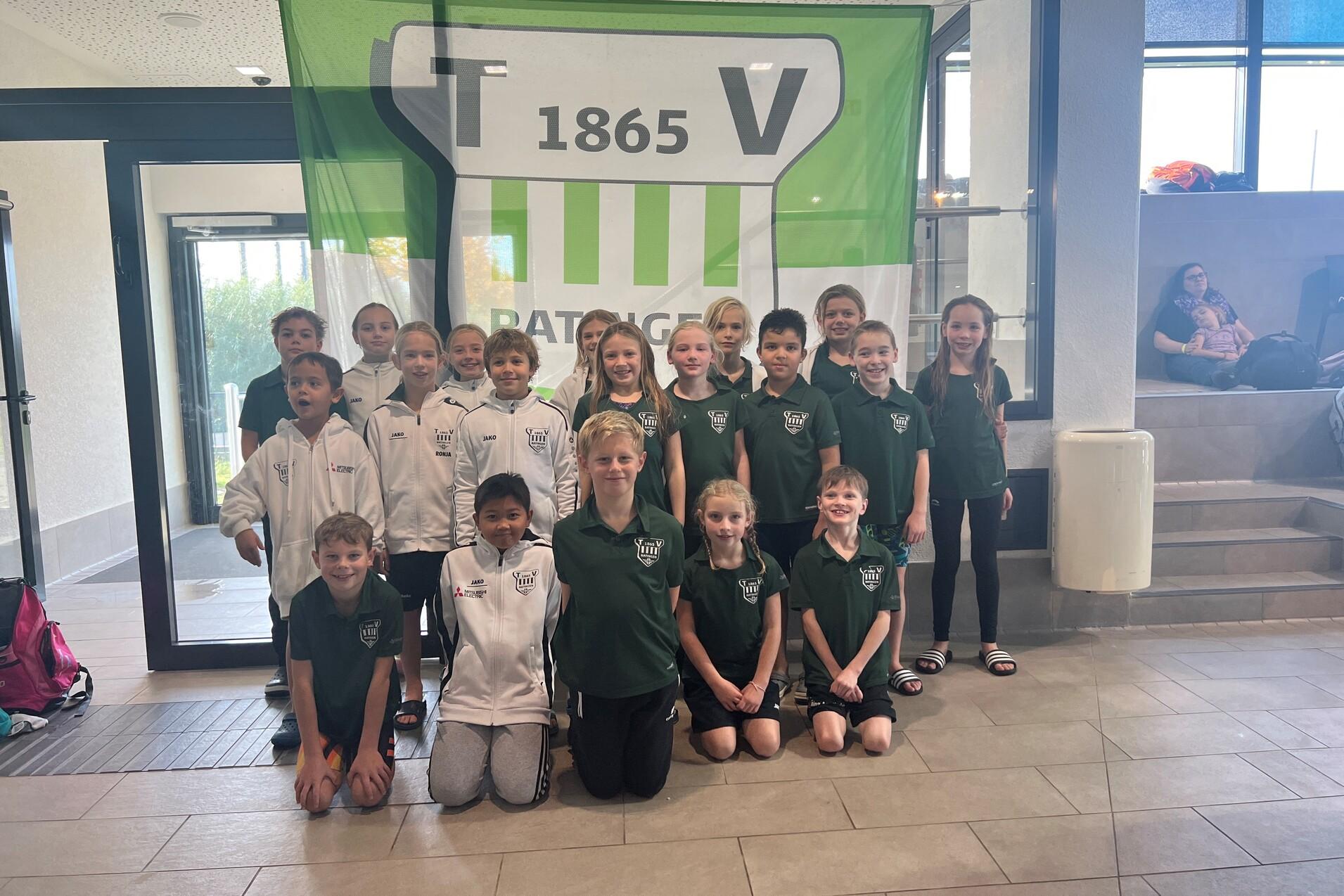 Gruppe von Kindern in Trainingskleidung, lächelnd vor einem großen Vereinsbanner in einem Foyer.