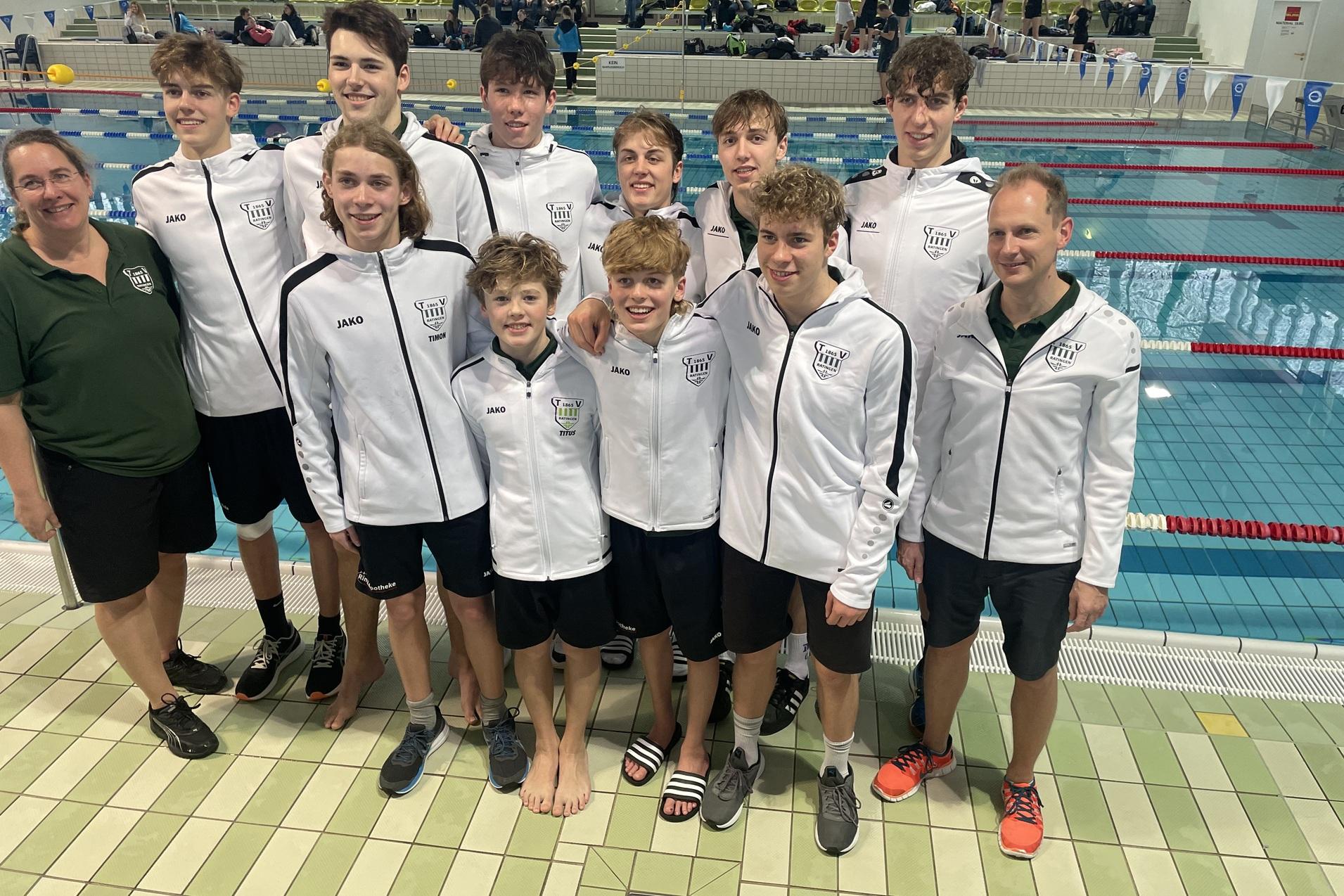 Gruppe junger Schwimmer in weißen Jacken posiert lächelnd vor einem Schwimmbecken. Ein Trainer steht ebenfalls dabei.